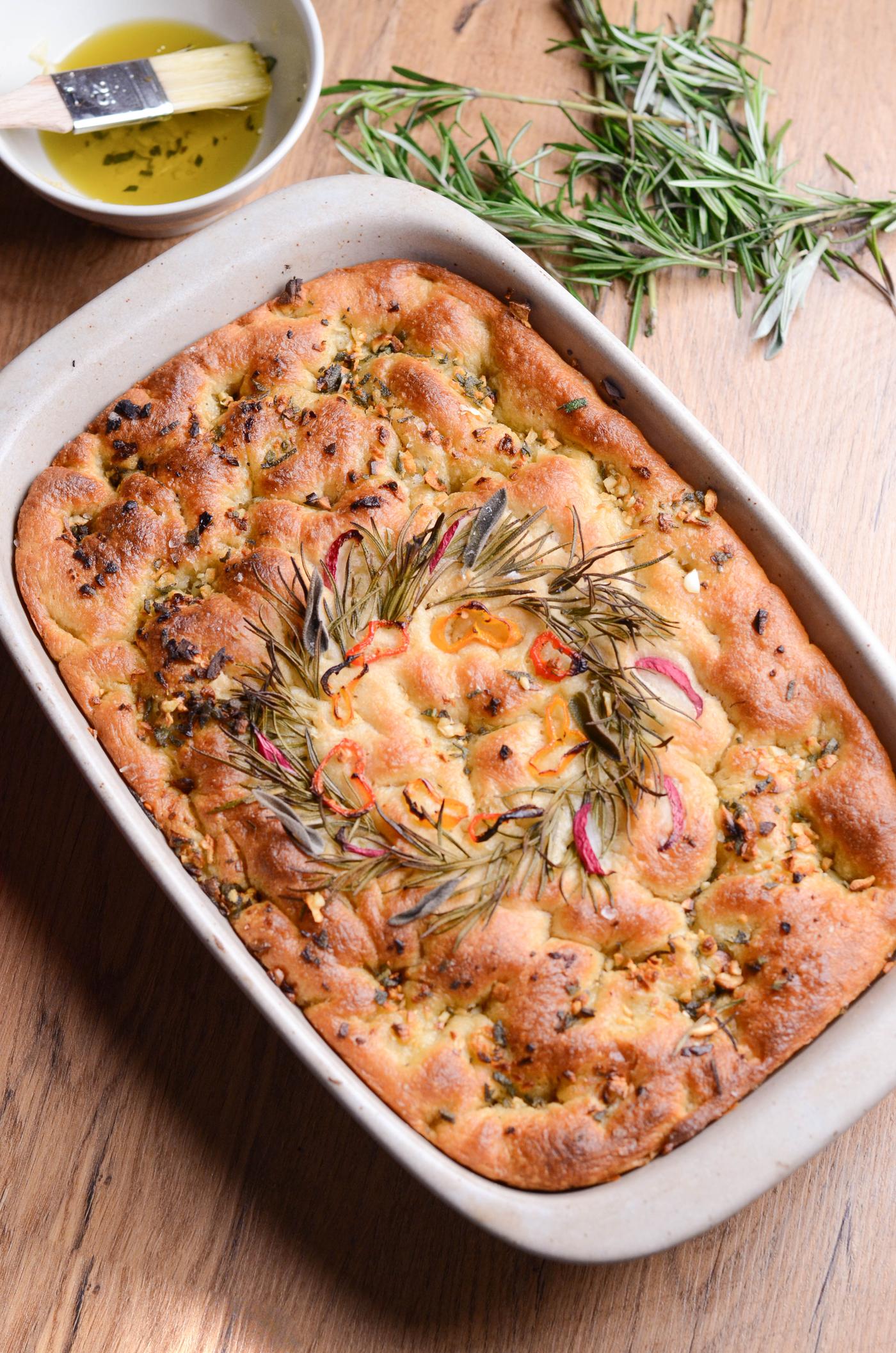Focaccia fraîchement cuite dans un plat rectangulaire, garnie d'une couronne de romarin et de tomates. Un plat contenant de l'huile d'olive et du beurre est posé à proximité sur une table en bois.