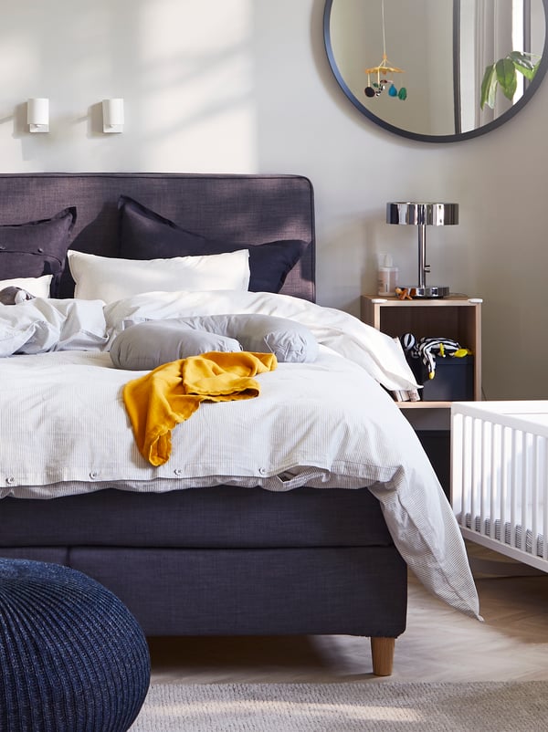 A bedroom with a DUNVIK dark-grey divan bed, grey/stripe quilt cover and pillow cases, two white reading lamps and a cradle.