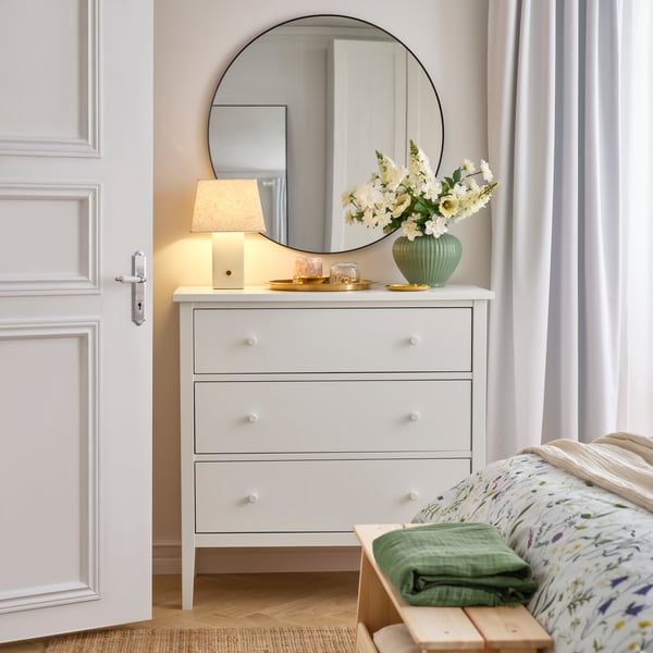 Ein Schlafzimmer mit rundem Spiegel über einer GULLABERG‑Kommode mit drei Schubladen. Auf der Kommode stehen eine Tischlampe und Blumen.