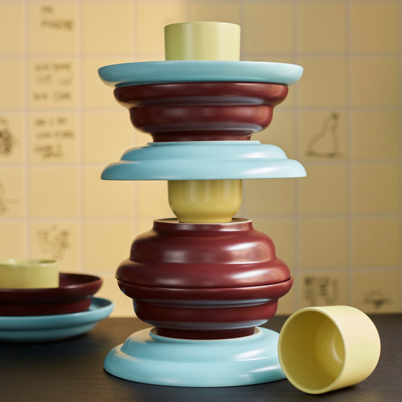 A SPJUTLILJA 3-piece serving set with dark purple bowls, yellow cups and light blue plates, in a tower on a dark table.