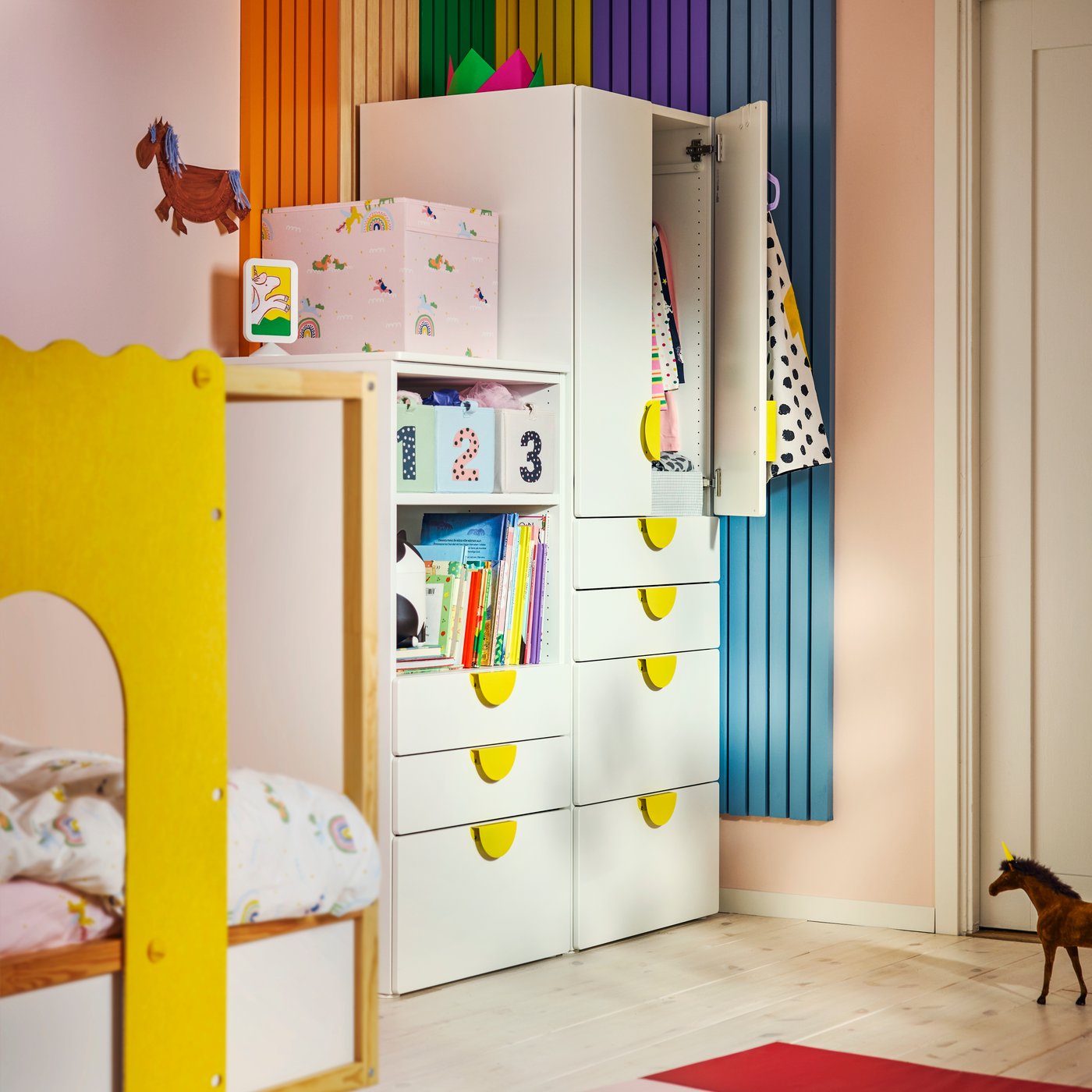 Ein Kleiderschrank der Serie „SMÅSTAD/PLATSA“ in Weiß mit gelben Griffen und offenen Regalböden in einem farbenfrohen Kinderzimmer mit gestreiften Wänden.