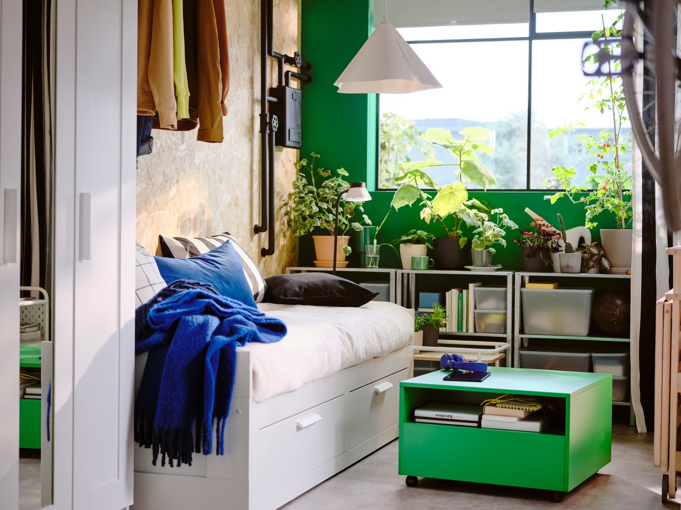 Bedroom with white day-bed, a bright green table on wheels, a window and shelves with lots of green plants in the background.