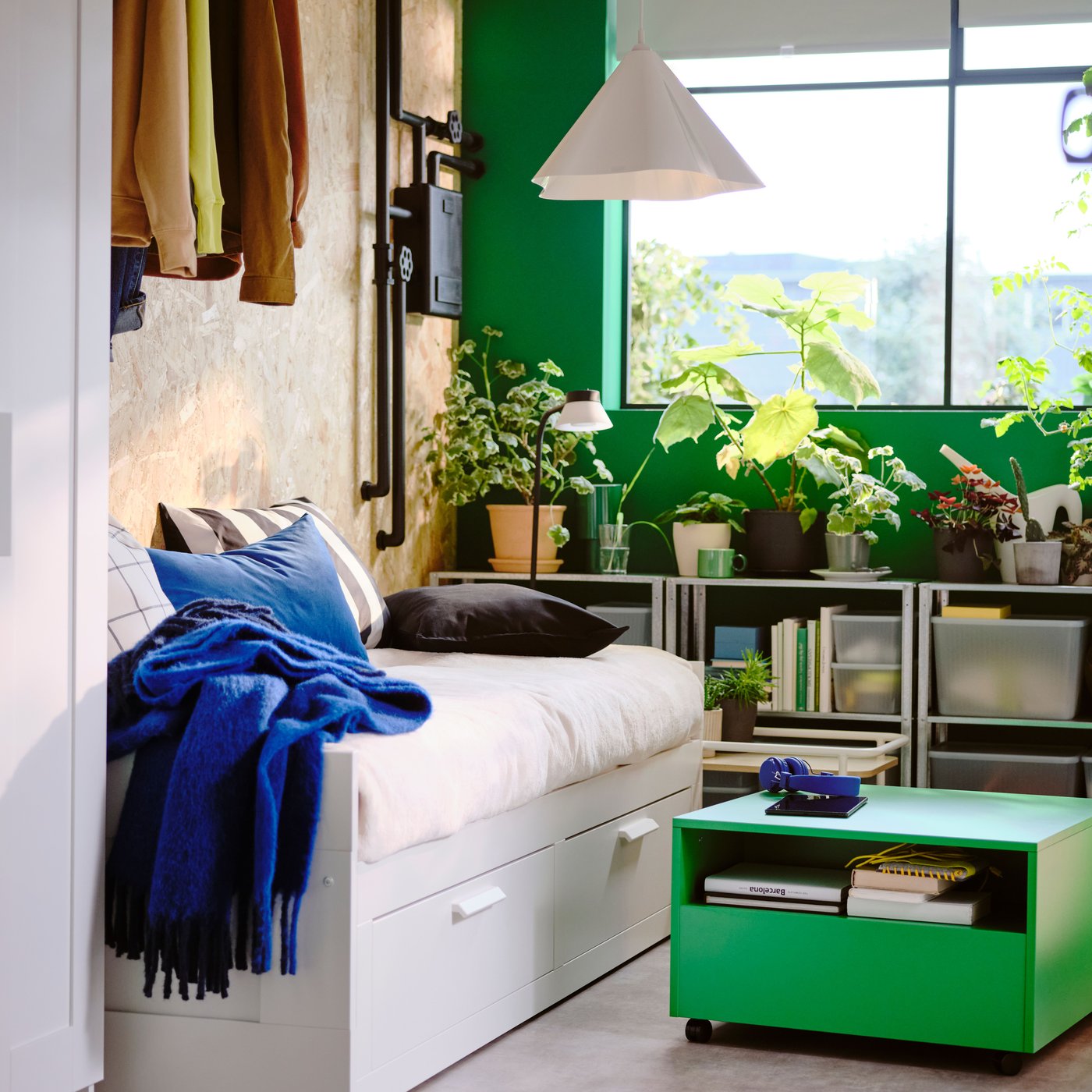 Bedroom with white day-bed, a bright green table on wheels, a window and shelves with lots of green plants in the background.