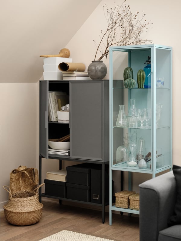 A RUDSTA glass door cabinet filled with glasses and other decorative objects beside a grey cabinet with an open door.