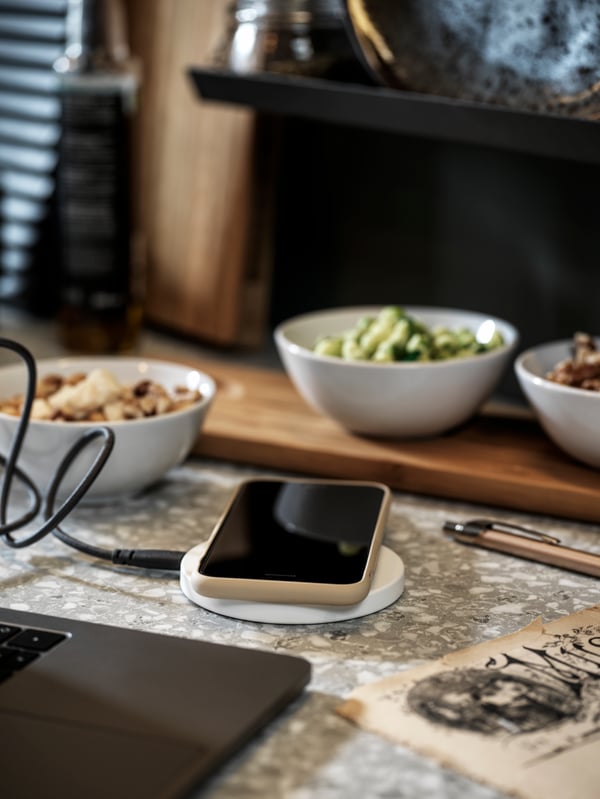 A mobile phone lies on top of a white LIVBOJ wireless charger on a kitchen countertop between a laptop and bowls of food.