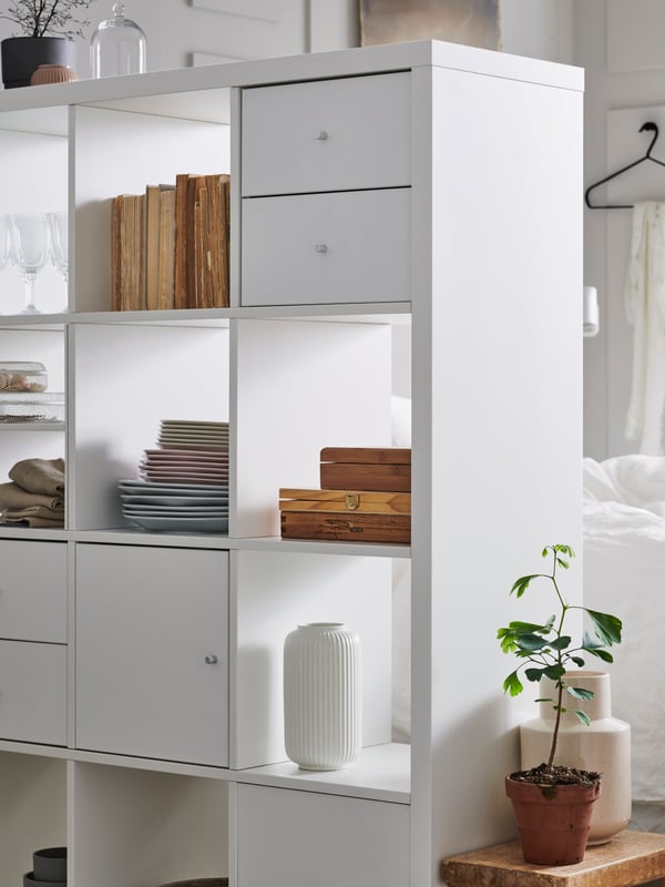 Part of a room-dividing white KALLAX shelving unit holding books and decorative items, and with both door and drawer inserts.
