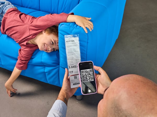 A boy is lying on a blue sofa in an IKEA store and laughing whilst his father scans the price tag using the IKEA app. 