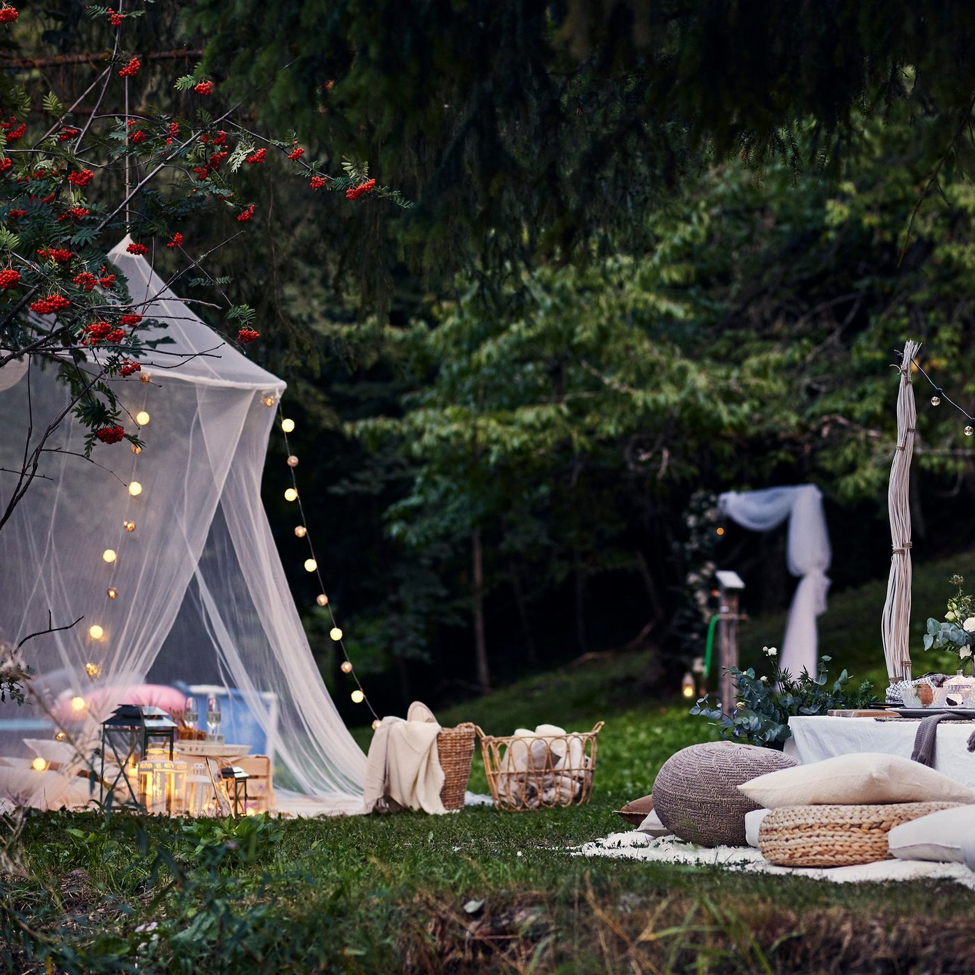 In einem Garten steht eine lange Tafel mit Tischdecke darüber, darum verschiedene Sitzkissen und Poufs. In den Bäumen hängen Laternen und Lichterketten als Dekoration