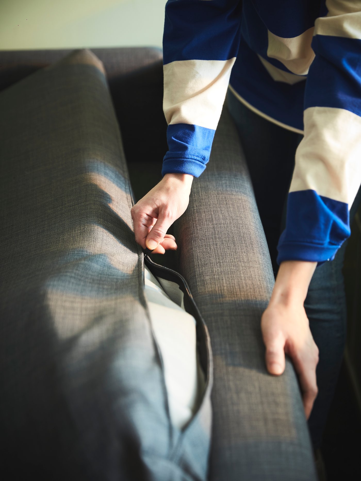 A person pulls the zipper of a dark-grey KLAGSHAMN back-cushion cover of a dark-grey FRIHETEN corner sofa-bed.