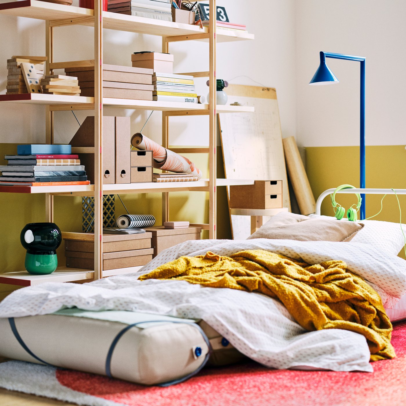 Bela IKEA PS 2026 fotelja na razvlačenje nameštena kao krevet s narandžastim tankim ćebetom, iza koje se nalaze drvene police na kojima su naslagane knjige i kutije.