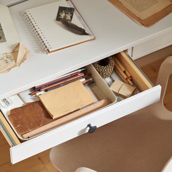 مكتب مع درجين HEMNES مطلي بالأبيض، وأحد الأدراج مفتوح وبداخله منظم بلاستيك/بيج وأدوات مكتبية.