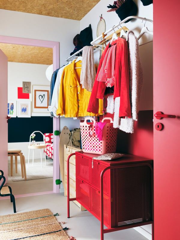 A colourful hallway with garments on white MULIG clothes bars on the wall above a red NIKKEBY chest of drawers.