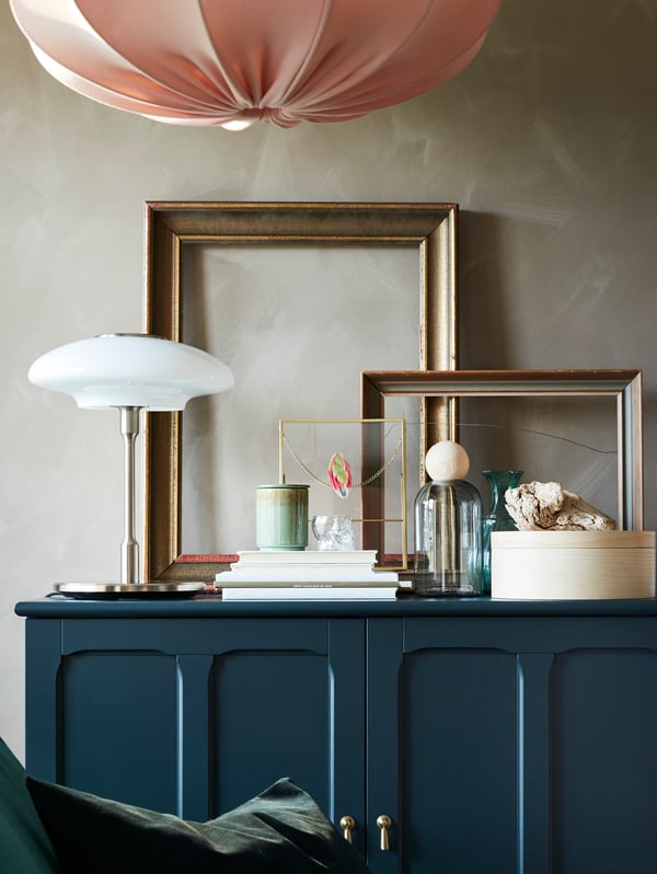Cadres dorés, livres, éléments décoratifs et une lampe de table nickelée/verre opalin sur une armoire bleu-vert.