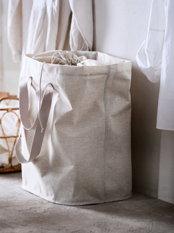 Una bolsa de ropa PURRPINGLA beige llena de textiles blancos sobre un piso de alfombra beige, entre dos prendas de ropa blanca colgada.