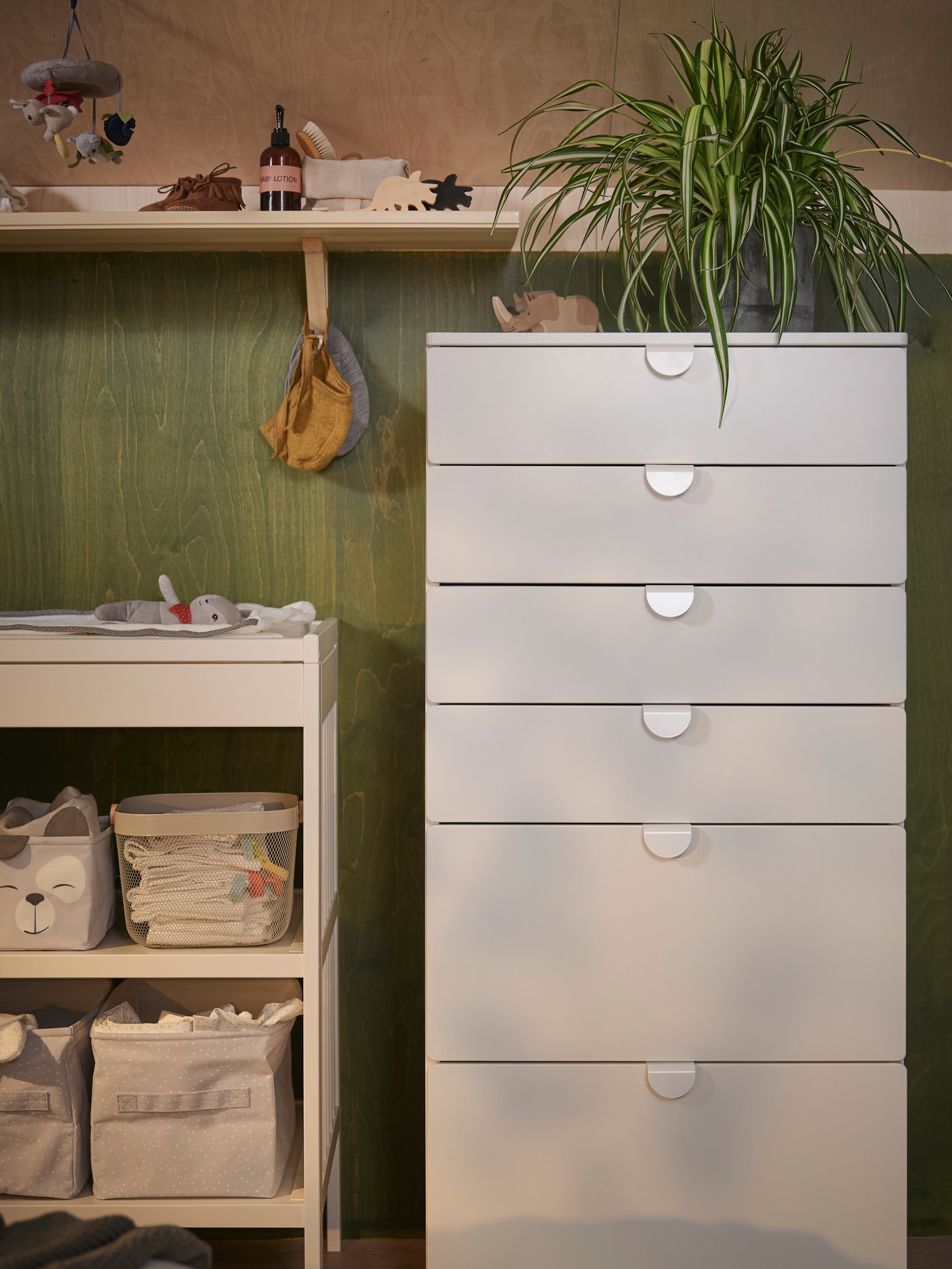 A white GULLIVER changing table by a white SMÅSTAD/UPPFÖRA chest with 6 drawers with a plant on top and a shelf above.