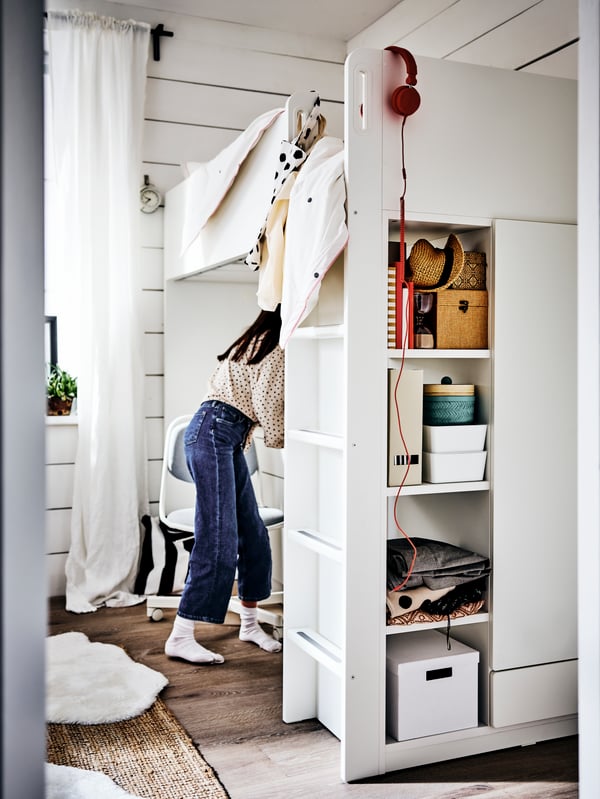 Une fille debout devant une chaise ÖRFJÄLL devant son bureau sous un lit mezzanine blanc. Des boîtes de rangement sont sur les étagères du cadre de lit.