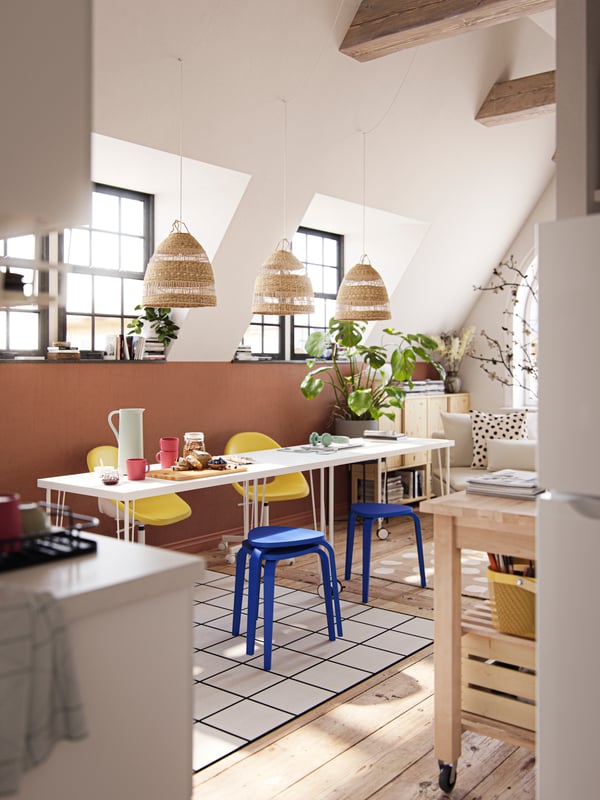 A sun-drenched dining area with two tables, blue stools, yellow chairs and three pendants made from natural materials.