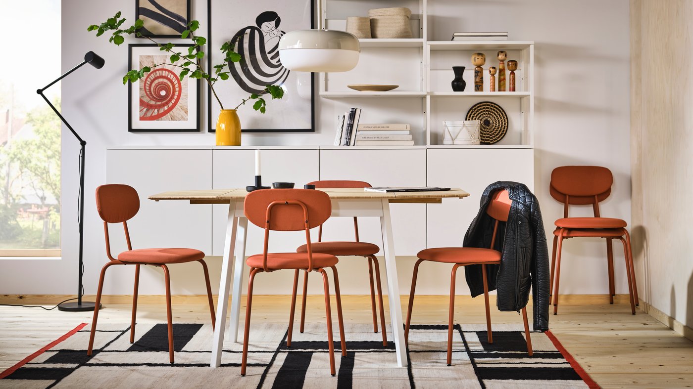 Es ist ein heller Essbereich mit gerahmter Kunst, einem PLATSA Regal und rotbraunen ÖSTANÖ Stühlen zu sehen, die rund um einen IKEA PS Klapptisch stehen.