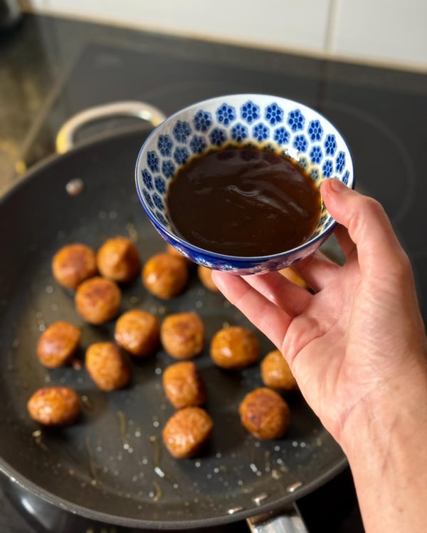 Une main tient un bol de sauce au-dessus d'une poêle remplie de boulettes de viande dorées qui cuisent sur la cuisinière.