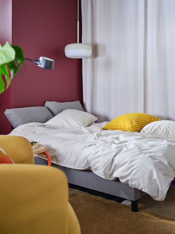 Sivi ÄLVDALEN sofa ležaj transformisan u gostinski krevet. Bela posteljina, lampe, žuti jastuk i bele zavese iza njega.