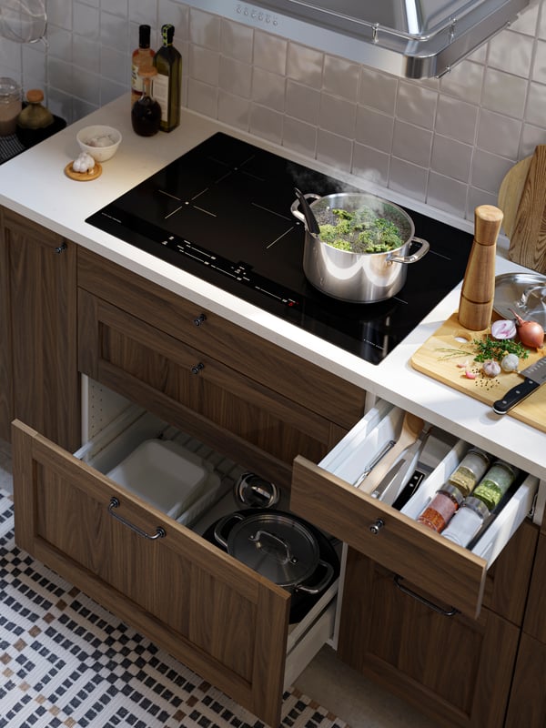 An IKEA 365+ pot with lid on a black induction cooktop, plus open kitchen drawers with pots, oven dishes and spices inside.