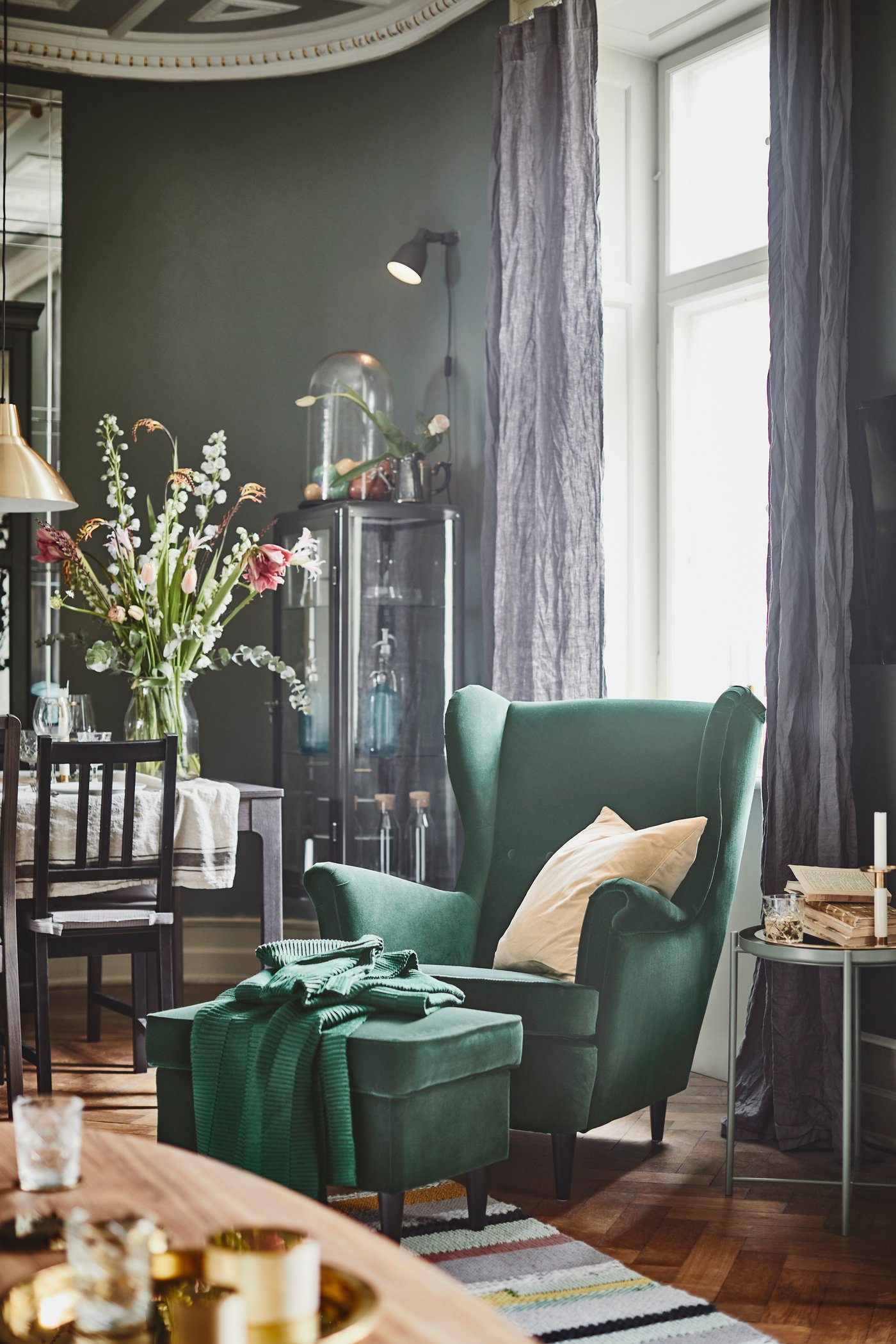 Un fauteuil à oreilles en velours vert avec un tabouret adapté est placé devant une fenêtre avec des rideaux gris foncé en lin et une table de salle à manger sur laquelle est placé un grand vase avec des fleurs 