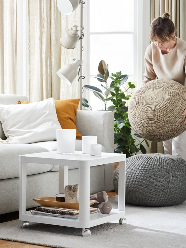 Woman in a living room with sofa, wheeled side table, rug and round pouffe. She’s holding a second pouffe, moving it.