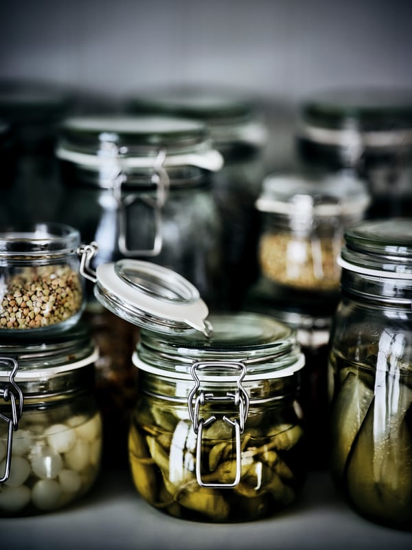 Une tablette sur laquelle sont rangés des bocaux KORKEN de différentes tailles remplis de marinades, de légumes et d’épices. Le couvercle d’un bocal à l’avant est ouvert.