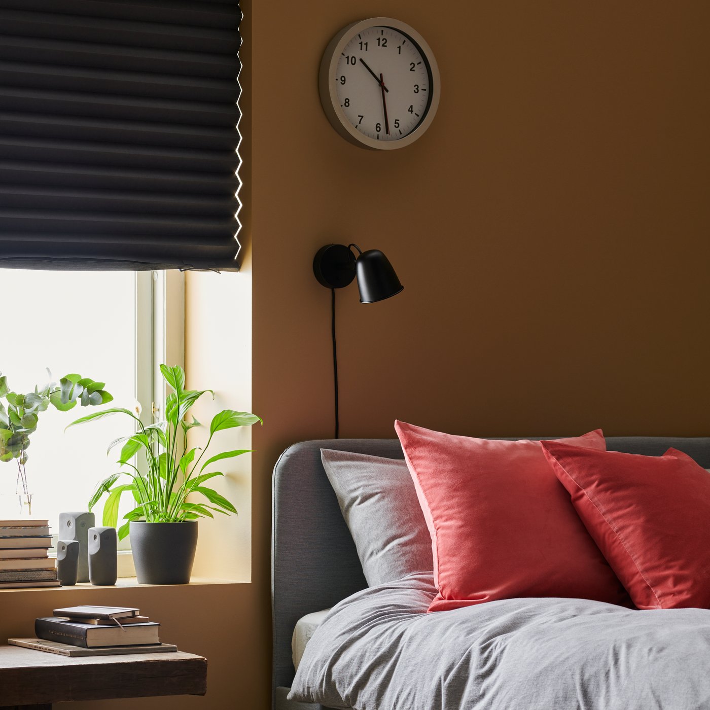 Vista de una habitación con paredes color tierra, una ventana con una cortina roller SCHOTTIS a medio bajar. En el borde de la ventana hay plantas y accesorios decorativos. Al costado de la ventana hay una cama con textiles IKEA