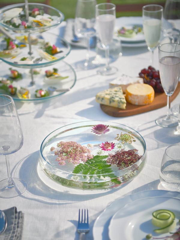 Blumen schwimmen in einer mit Wasser gefüllten BERÄKNA Servierschale, die auf einem Tisch steht, der mit SVALKA Gläsern und einer KVITTERA Etagere eingedeckt ist.