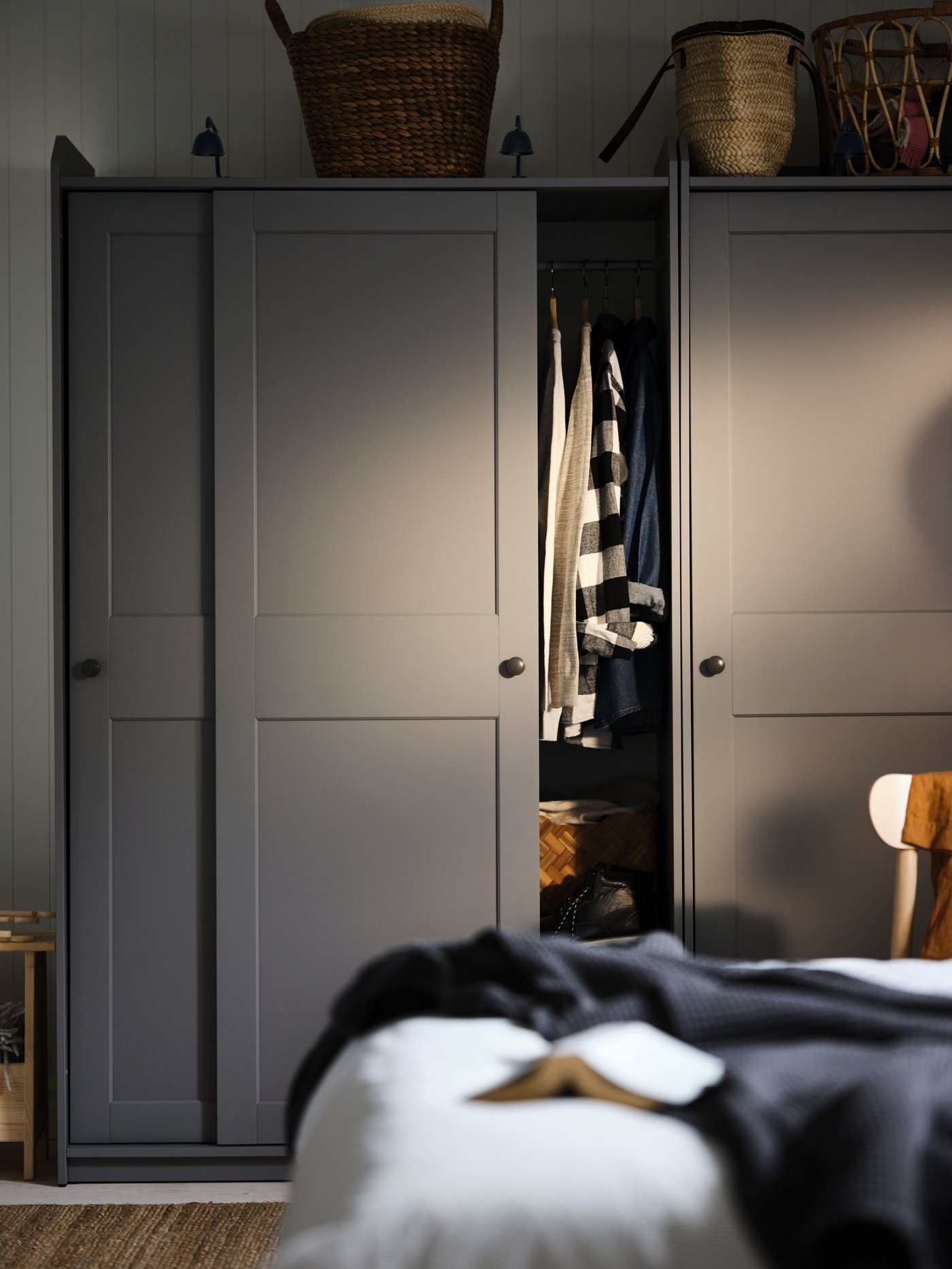 In a bedroom a grey HAUGA wardrobe with sliding doors and a few storage baskets on top of it. A chair in front of it.