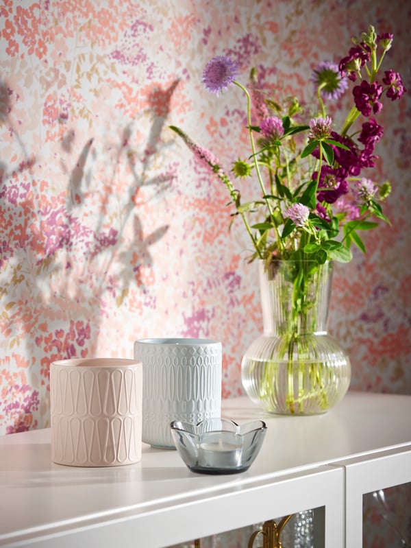 Des bougies parfumées PÅKOSTAD dans leurs pots en céramique, un photophore VANLIGEN gris et un vase en verre PÅDRAG contenant des fleurs sur une armoire blanche à portes vitrées.