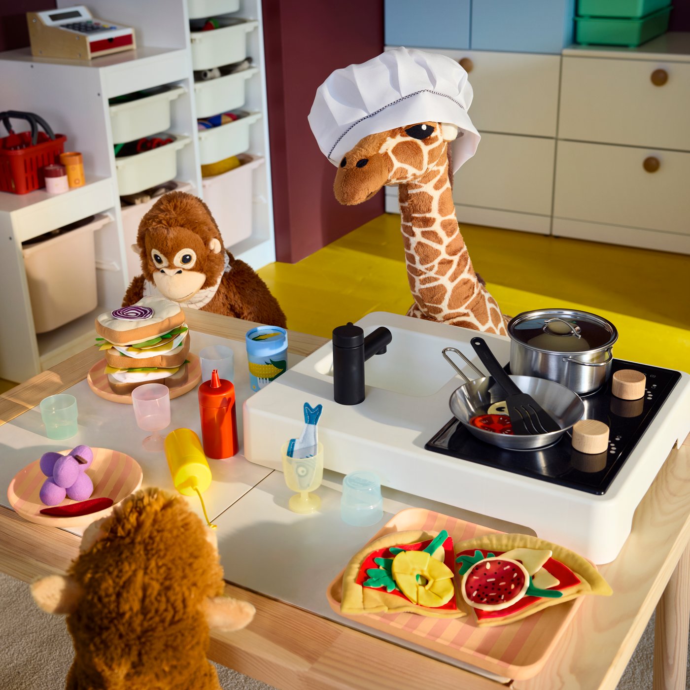 A plush giraffe in a chef hat and orangutans play with the STENMUS mini-toy kitchen and toy street food in a child’s room.