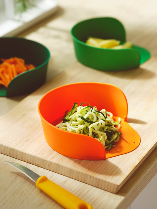 An APTITLIG chopping board in bamboo, three UPPFYLLD preparation bowls in mixed colours and a yellow UPPFYLLD paring knife.