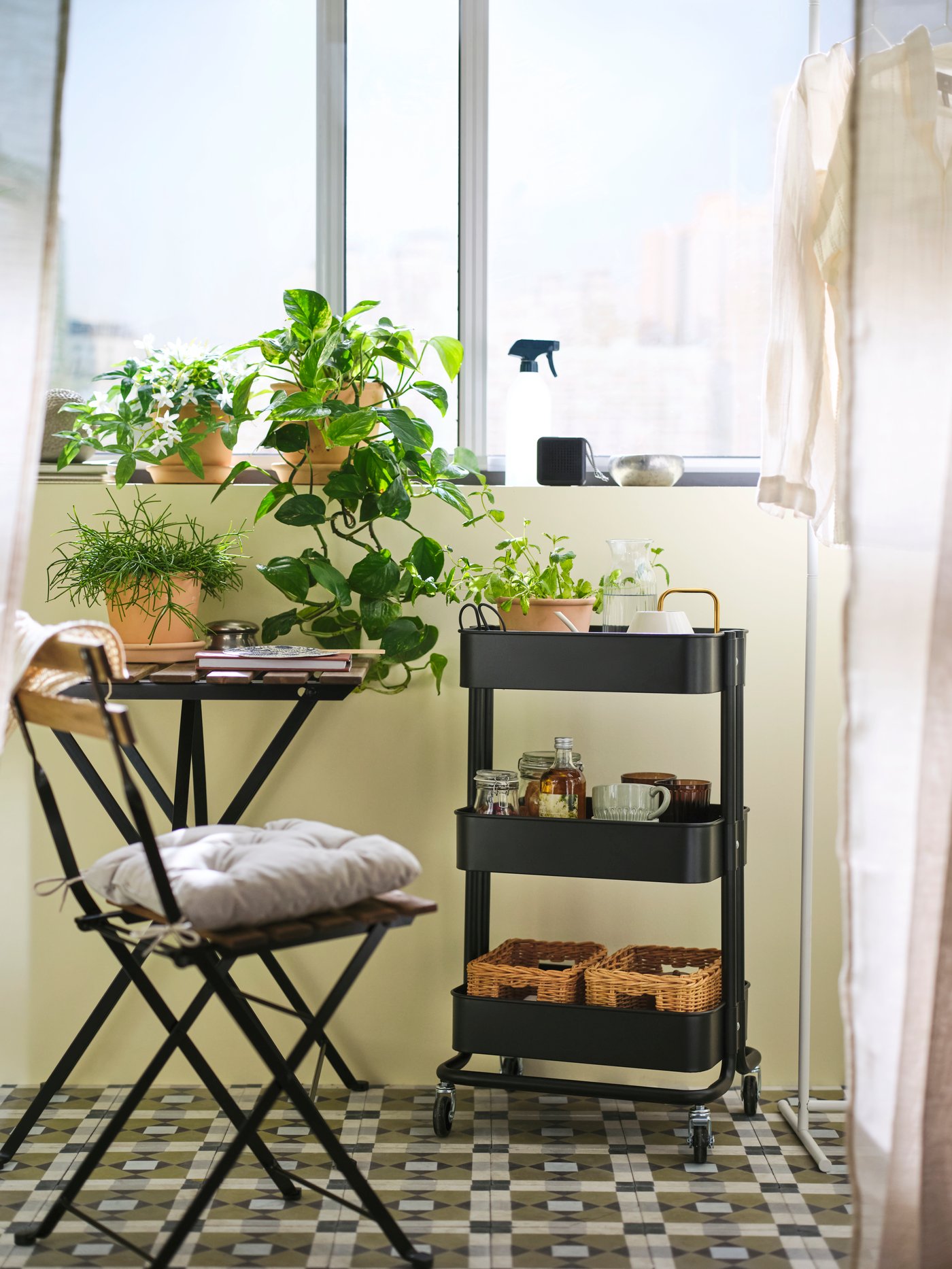 A black, coated-steel RÅSKOG trolley holding potted herbs, jars, glassware and baskets is in an enclosed balcony by a table.