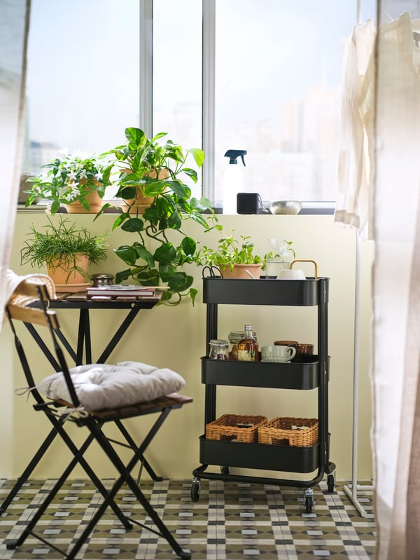A black, coated-steel RÅSKOG trolley holding potted herbs, jars, glassware and baskets is in an enclosed balcony by a table.