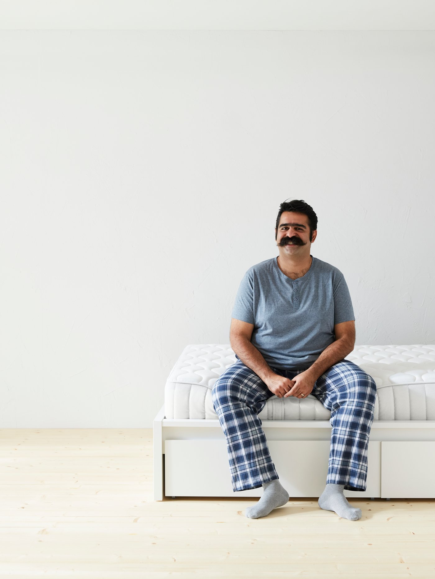 A man with a moustache and wearing a greyish-blue t-shirt and blue plaid pyjama pants sits on the edge of a mattress.