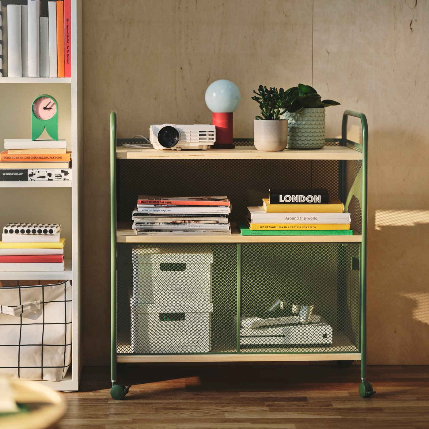 A living room with a dark green NÅLMAKARE cabinet on castors, displaying books and a plant on a shelf.