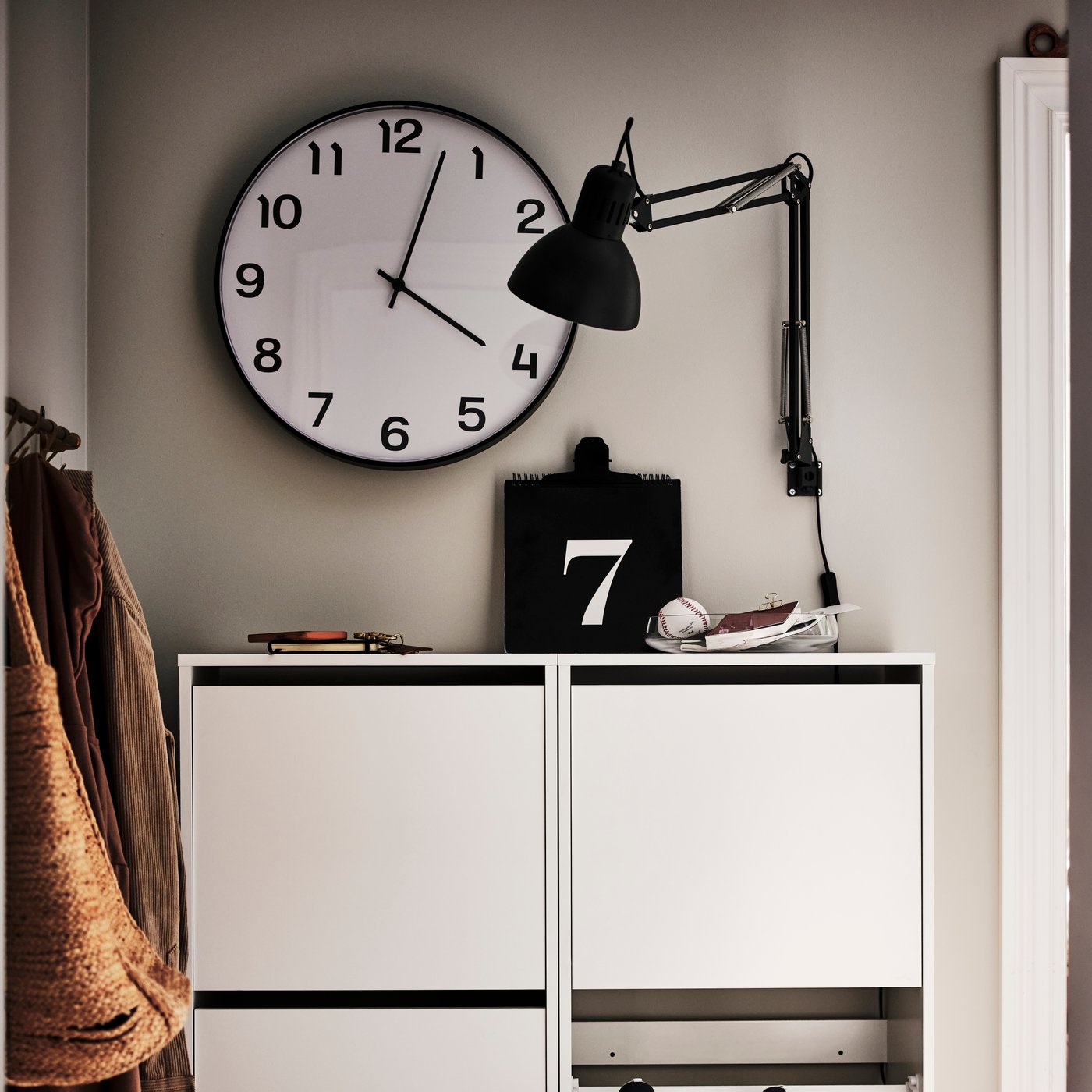 Une horloge murale PLUTTIS noire suspendue sur un mur au-dessus d’un meuble de rangement blanc. 