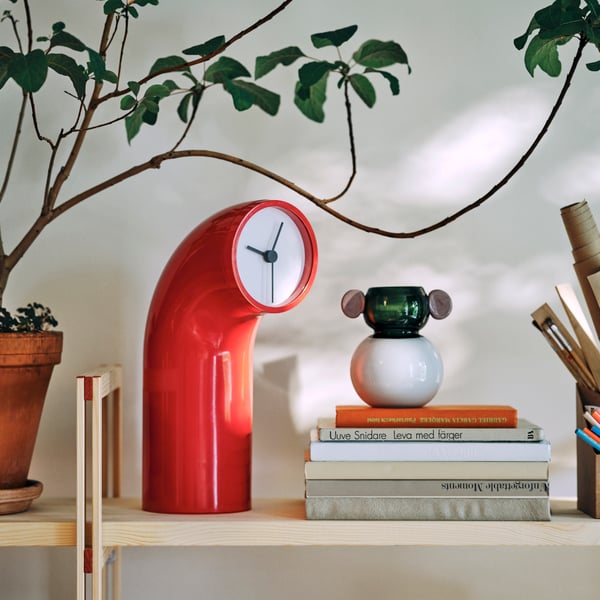 Close-up of a red sculptural IKEA PS 2026 table clock placed next to stacked books.