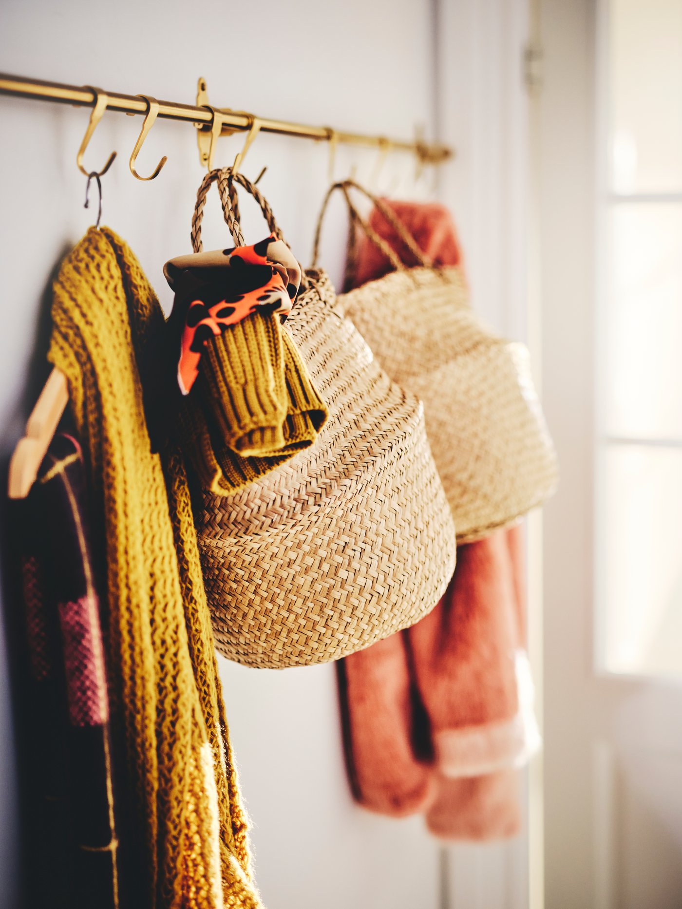 Two FLÅDIS baskets stuffed with clothing items and two scarfs on a hanger, hanging on gold-tinted hooks.