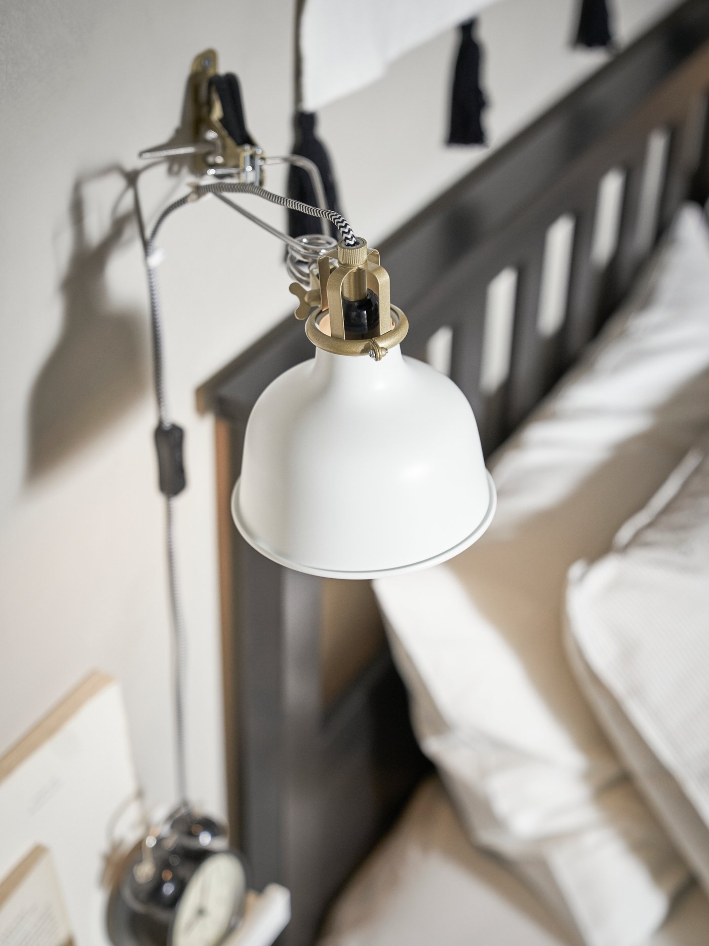 A RANARP wall/clamp spotlight in off-white is mounted on a bright grey wall next to a dark bed with white bed linen.
