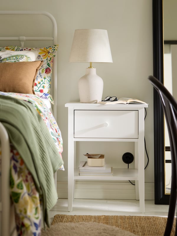 Una mesa de noche GULLABERG blanca con una lámpara de mesa BILDVÄDER en color hueso, junto a una cama con ropa de cama floral.