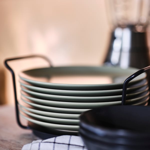 A stack of eight 26 cm matt green FÄRGKLAR plates on a wooden surface with a hint of a tea towel in the foreground.
