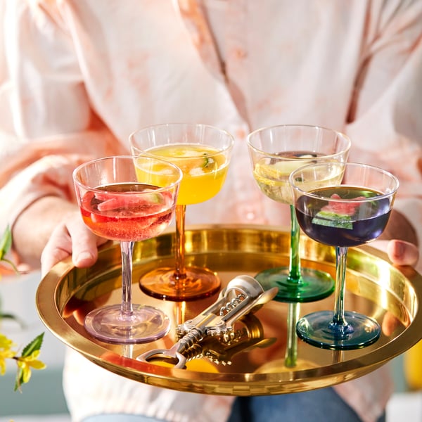 Vier TJÄRLEK Champagnerschalen mit unterschieldich farbigen Stielen stehen auf einem warm schmimmernden, goldenen Tablett.