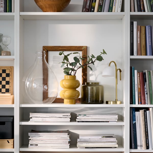 Netjes georganiseerde witte BILLY boekenkasten met een geel-beige PÄRLHÄGG vaas en een TVÅMASTAD draagbare lamp die een plank delen.