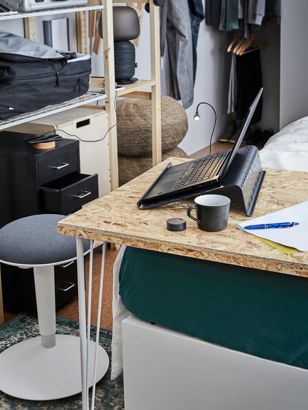 Ein Laptop steht auf einem schwarzen BRÄDA Laptophalter auf einem Tisch neben einem Becher, einem Stift und etwas Papier; im Hintergrund sind ein Hocker und offene Aufbewahrung zu sehen.