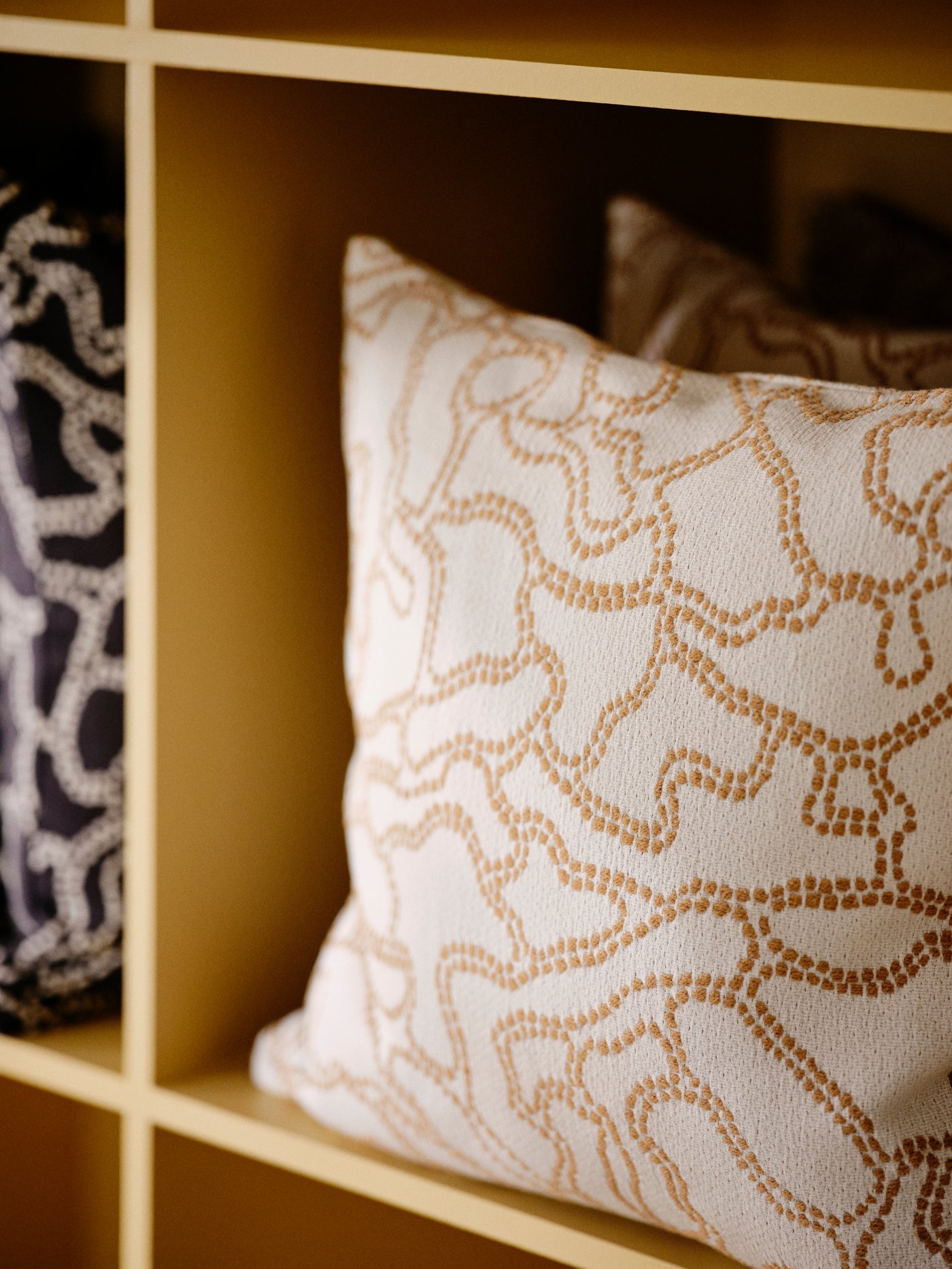 Una fodera per cuscino GULDFLY bianco sporco con lavorazione jacquard in giallo-beige dentro uno scaffale.