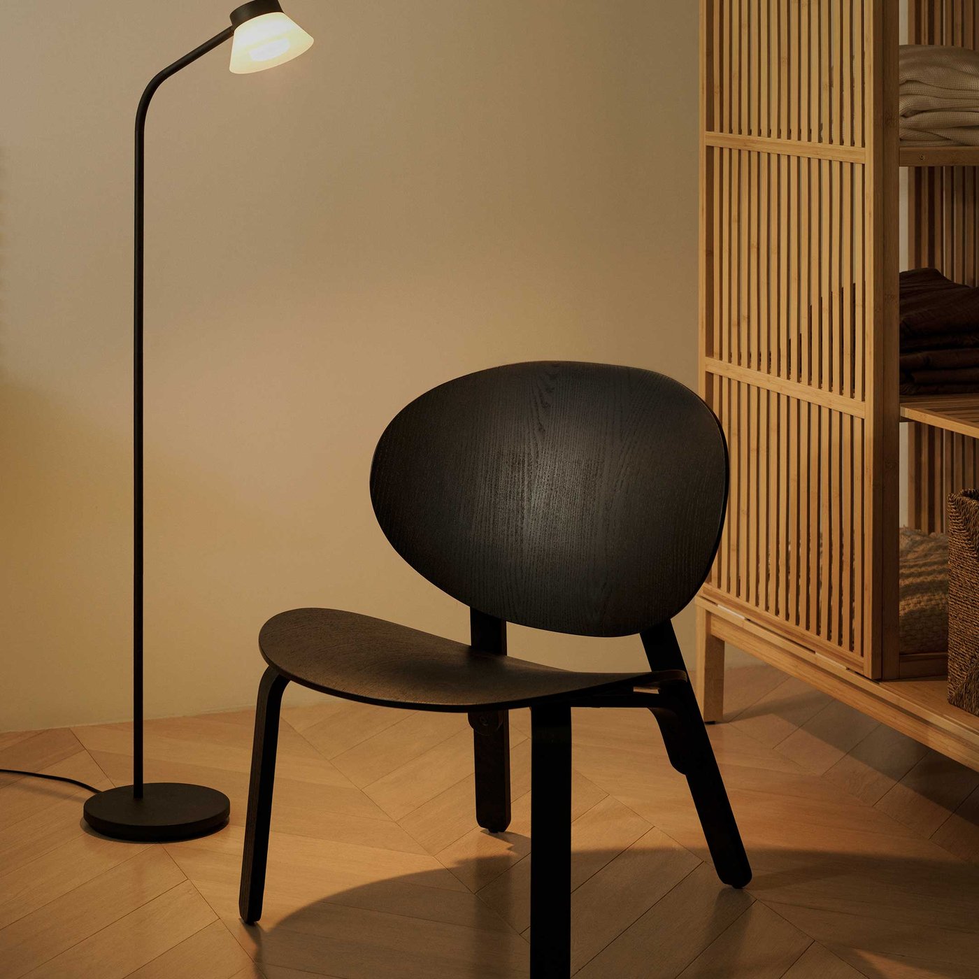 A stylish and functional corner, perfect for adding a touch of modern design to your home. A sleek black chair with a distinctive rounded back and seat takes centre stage, offering a comfortable spot for a moment of quiet. Next to it, a tall, slender black floor lamp with a white, conical shade casts a soft, inviting glow, ideal for reading or creating a cosy atmosphere. To the right, a natural wooden slatted shelving unit provides a practical and attractive storage solution, with neatly folded textiles visible on its shelves. The warm wooden flooring ties the space together, creating a harmonious and welcoming feel.