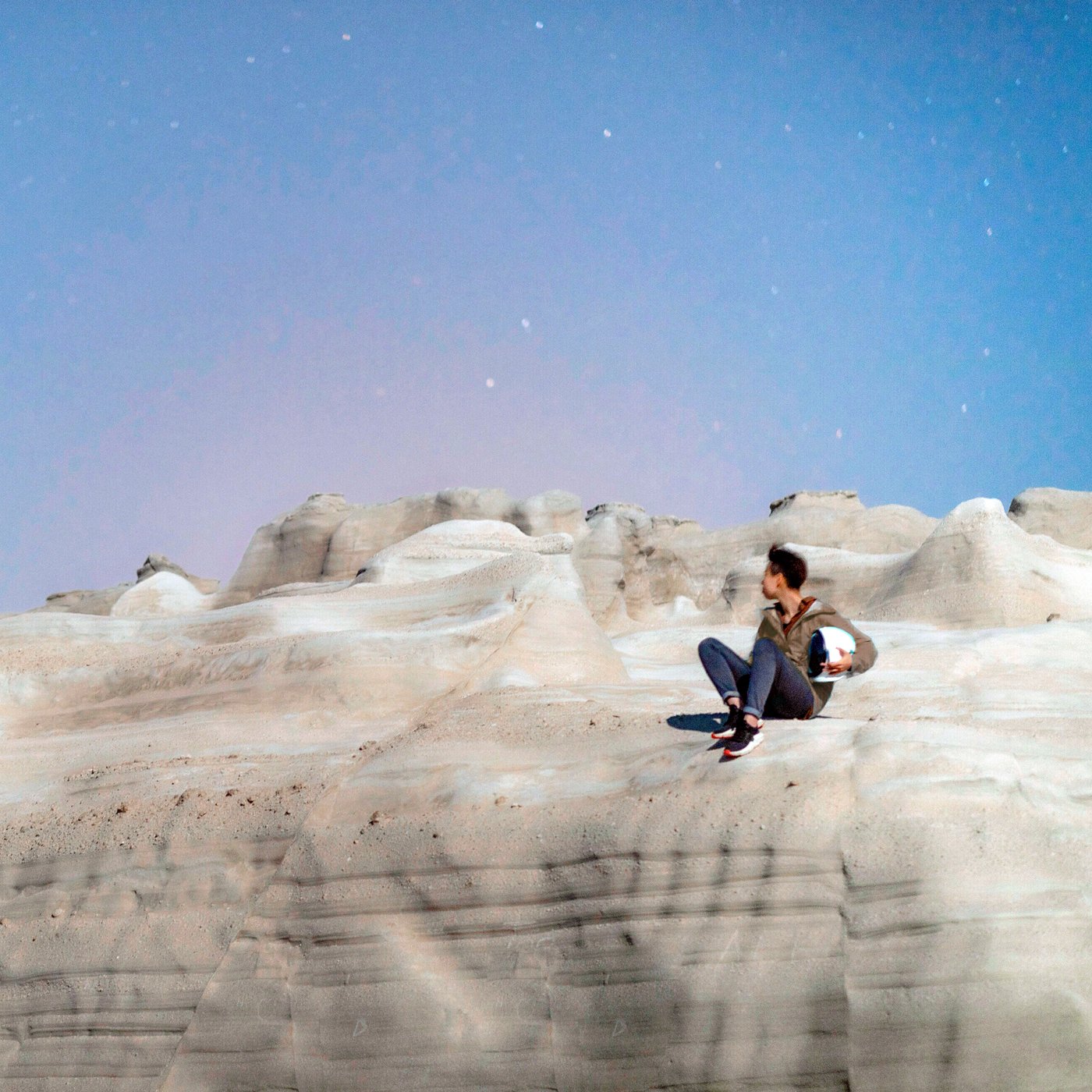 A woman sits outdoors on a big, smooth rock, with a blue sky behind her.